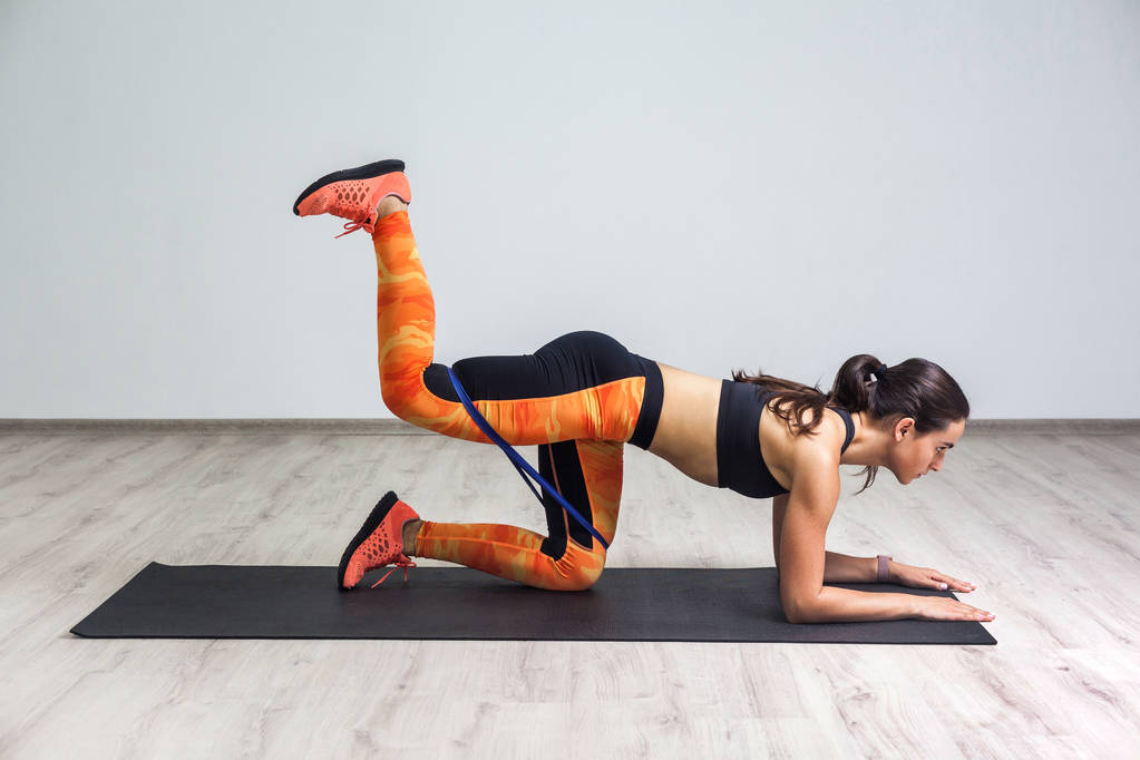 Vue latérale d'une jeune femme sportive forte et en forme en tenue de sport debout sur un genou sur un tapis avec jambe relevée et jambes d'entraînement et fessiers musculaires tout en utilisant une bande élastique de fitness dans la salle de gym
 - Photo, image
