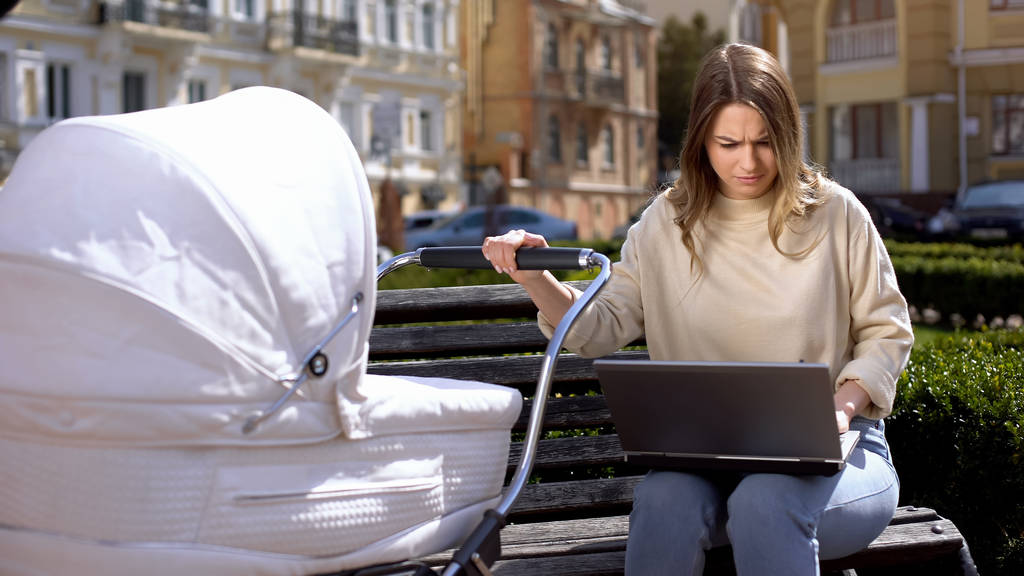 Occupé jeune femme essayant de travailler ordinateur portable et balançant nouveau-né dans landau, garde d'enfants
 - Photo, image