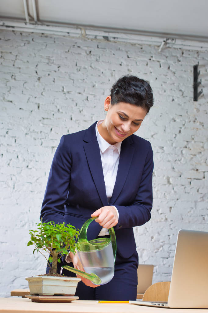 オフィスのレンガの壁に対して机の上に植物を水やり若いビジネスウーマンの笑顔 - 写真・画像
