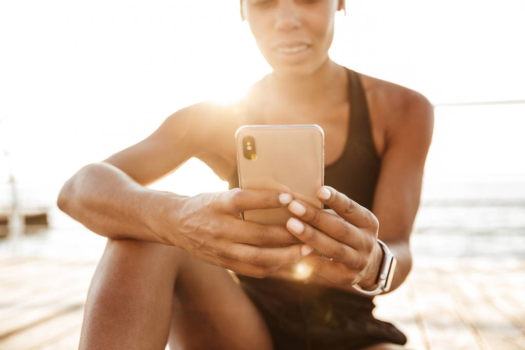 海のそばに座りながらスマートフォンを持つ日当たりの良い女性の写真 - 写真・画像