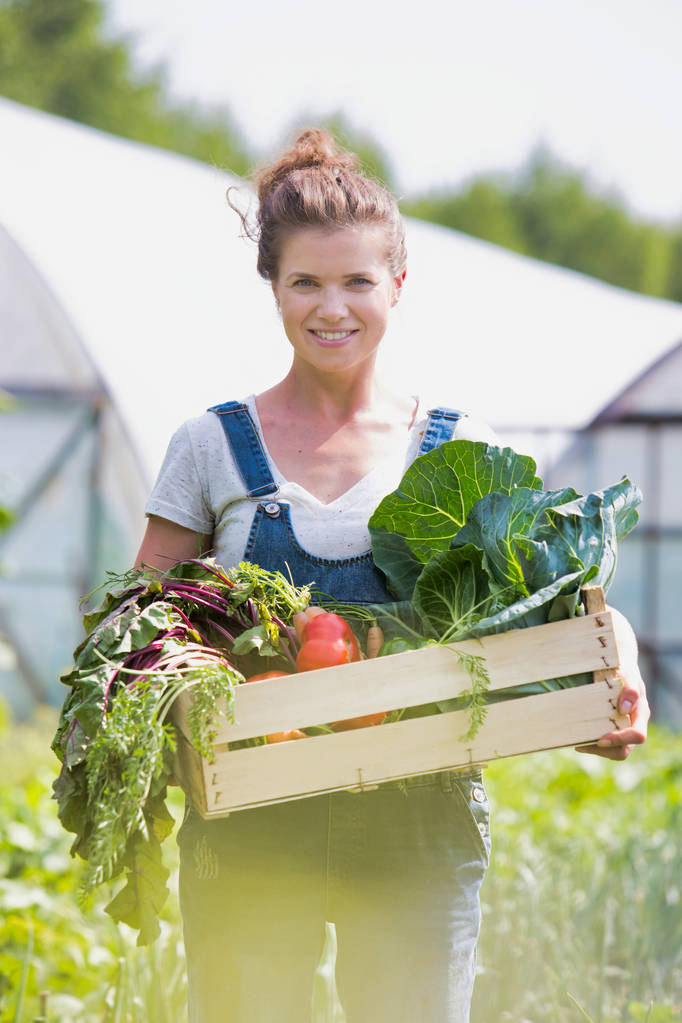 Porträt einer lächelnden erwachsenen Frau, die auf einem Bauernhof Gemüse in einer Kiste hält - Foto, Bild