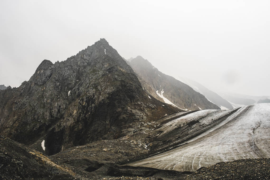 ein hoher peramidaler Berg in der Nähe eines schmelzenden Gletschers. Die felsigen Hänge sind mit Schnee bedeckt. volumetrisches Foto vom Altai. Scharfe Gipfel werden von undurchdringlichem Nebel verdeckt. der Beginn der Gebirgsflüsse. - Foto, Bild