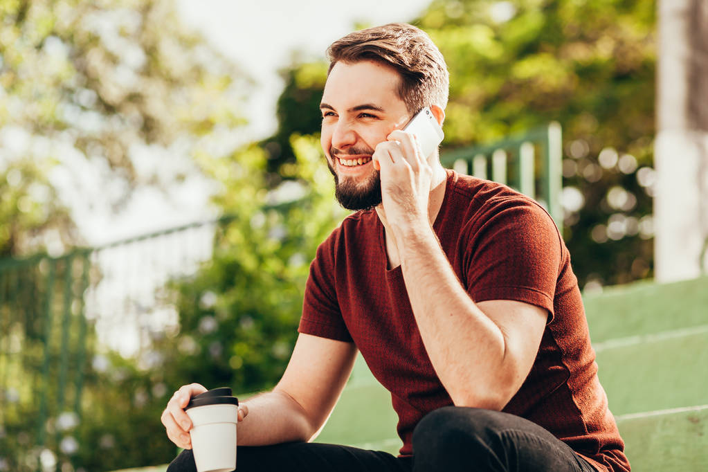 Mladý muž sedící ve veřejném parku pije kávu a mluví na  - Fotografie, Obrázek