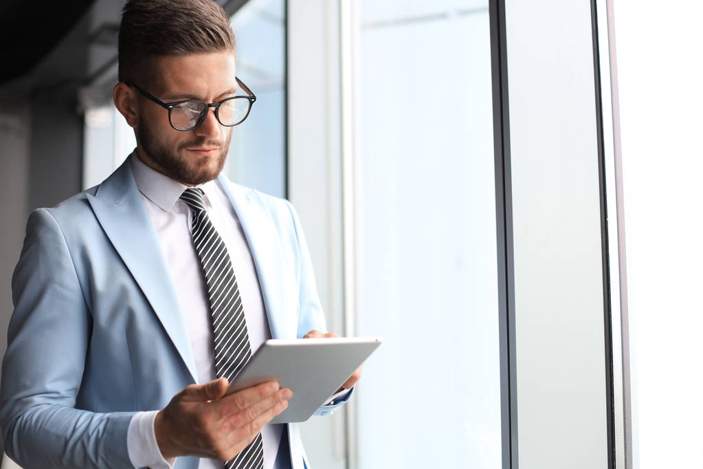 Moderno uomo d'affari in abbigliamento formale utilizzando tablet digitale mentre in piedi vicino alla finestra in ufficio
 - Foto, immagini