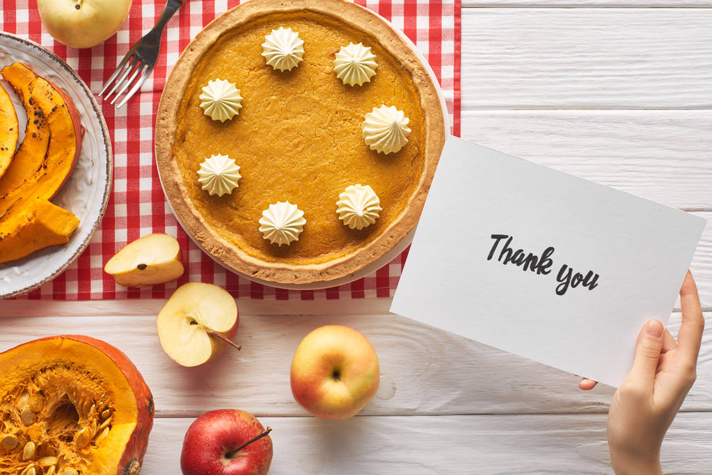 vista cortada da mulher segurando cartão de agradecimento perto de torta de abóbora na mesa branca de madeira com maçãs
 - Foto, Imagem
