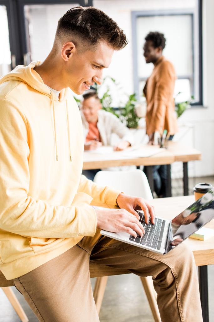 giovane uomo d'affari che utilizza laptop vicino a colleghi multiculturali che lavorano in ufficio
 - Foto, immagini