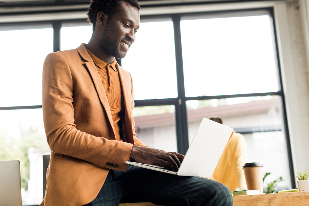 усміхнений афроамериканський бізнесмен сидить на столі і використовує ноутбук
 - Фото, зображення