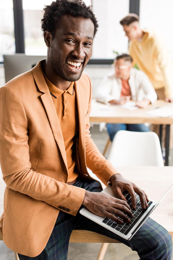 iloinen afrikkalainen amerikkalainen liikemies katselee kameraa istuessaan pöydällä ja käyttäen kannettavaa tietokonetta
 - Valokuva, kuva