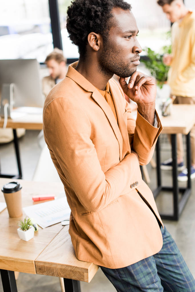 pensativo hombre de negocios afroamericano mirando hacia otro lado mientras está de pie en la oficina cerca de colegas
 - Foto, imagen