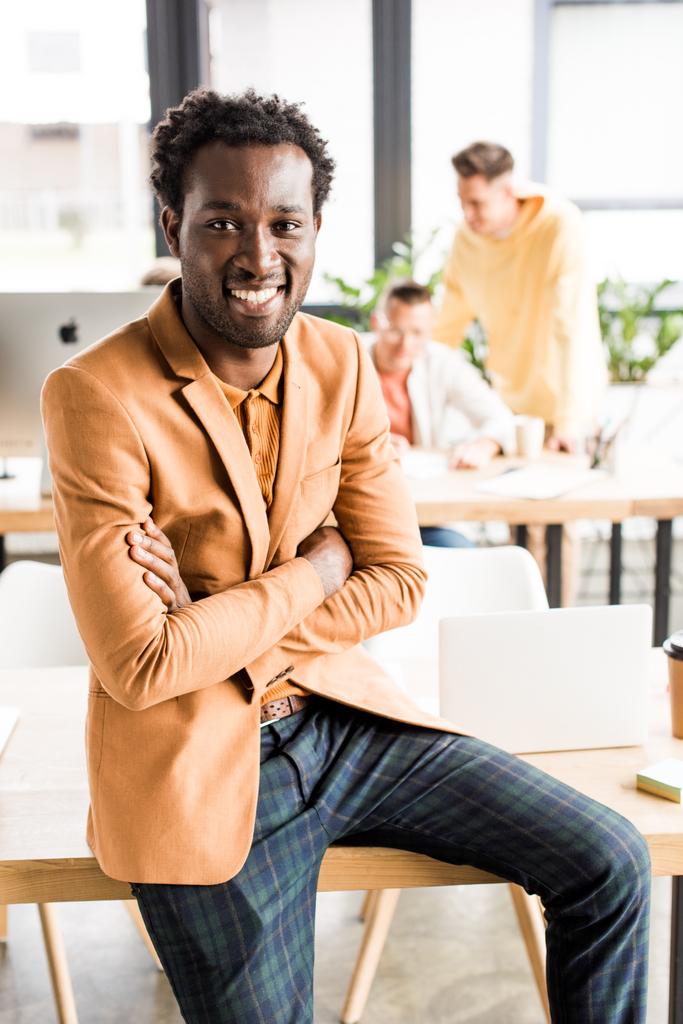 χαμογελαστός Αφροαμερικάνος επιχειρηματίας κάθεται στο γραφείο με σταυρωμένα χέρια - Φωτογραφία, εικόνα