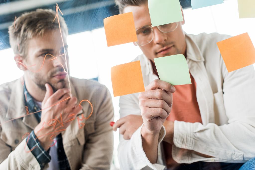 two young businessmen looking at sticky notes fixed on glass board in office - Photo, Image
