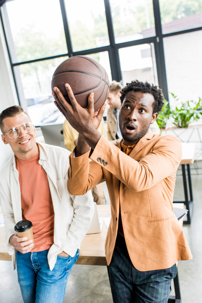 africký americký podnikatel hraje s volejbal, zatímco stojí poblíž kolega drží kávu jít do úřadu - Fotografie, Obrázek