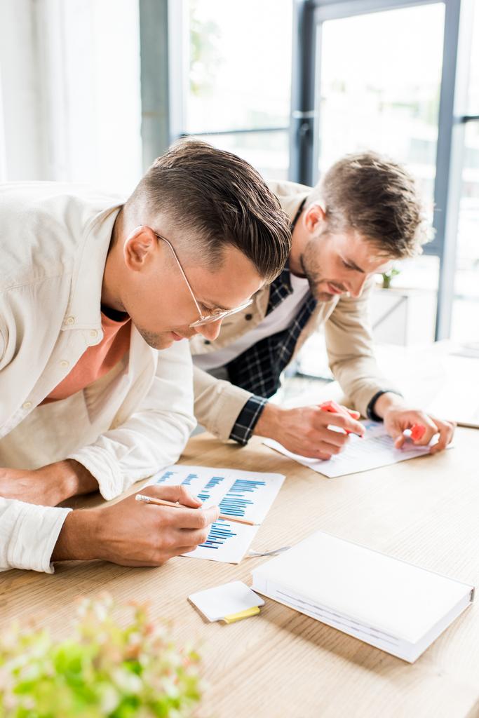 два молодых бизнесмена анализируют документы с графиками и диаграммами во время совместной работы над стартап-проектом в офисе
  - Фото, изображение