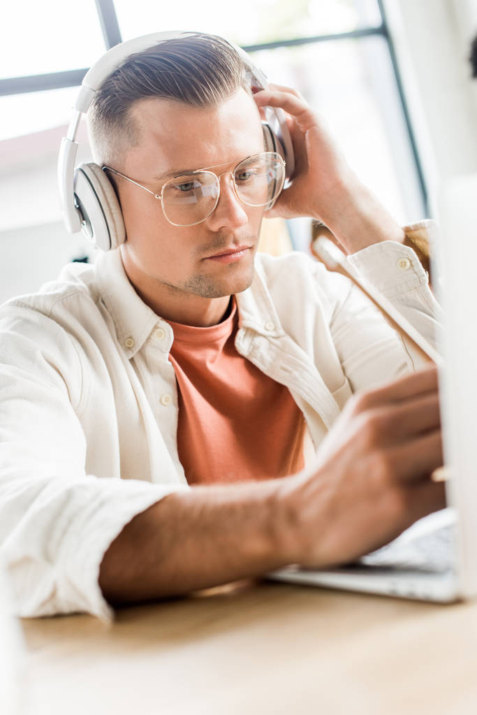 young, thoughtful businessman sitting in headphones and using laptop in office - Photo, Image