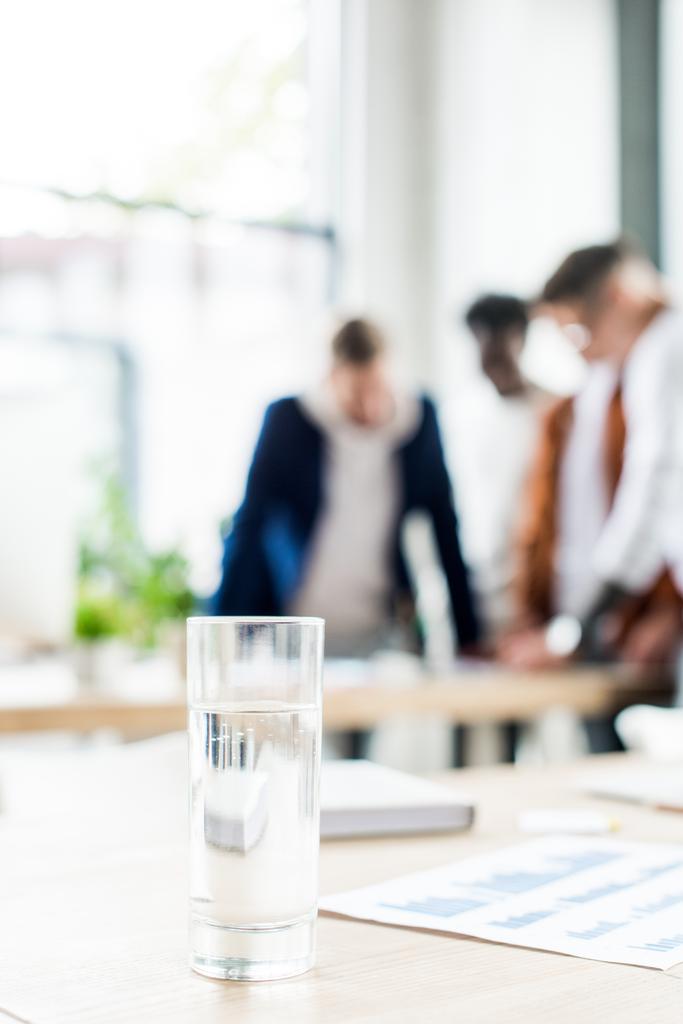 szelektív fókusz üveg vízzel az asztalon közel üzletemberek állnak a munkahelyen az irodában - Fotó, kép