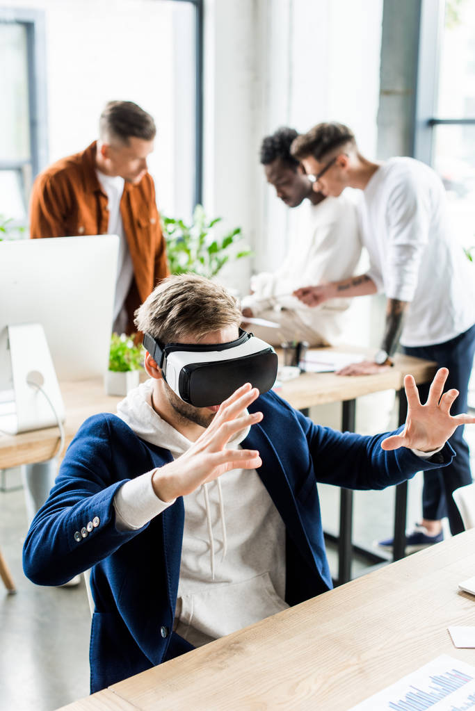 fiatal üzletember használ vr headset és gesztikuláció, miközben multikulturális kollégák dolgoznak az irodában - Fotó, kép