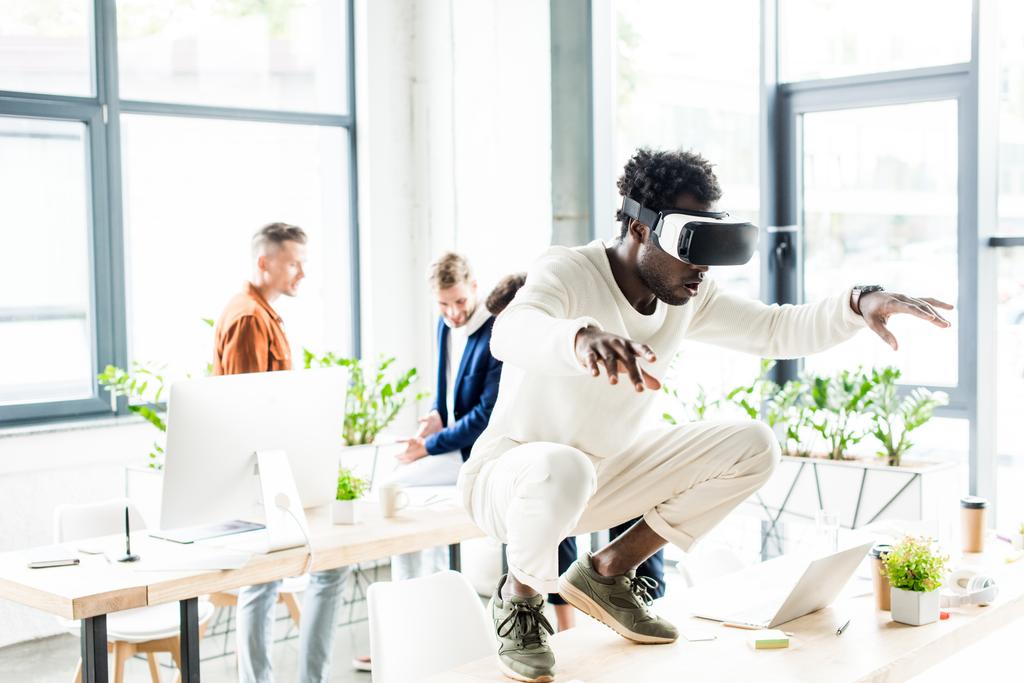 Afrykański amerykański biznesmen squatting na stole podczas korzystania z zestawu słuchawkowego VR, i współpracownicy pracujący w biurze - Zdjęcie, obraz