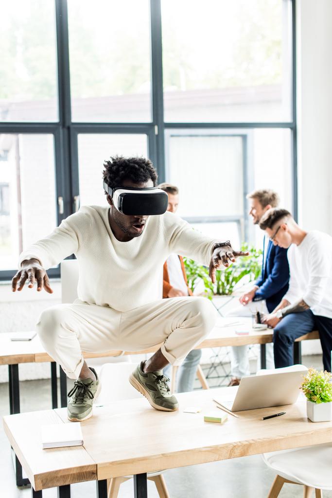 Afrykański amerykański biznesmen squatting na stole podczas korzystania z zestawu słuchawkowego VR, i współpracownicy pracujący w biurze - Zdjęcie, obraz