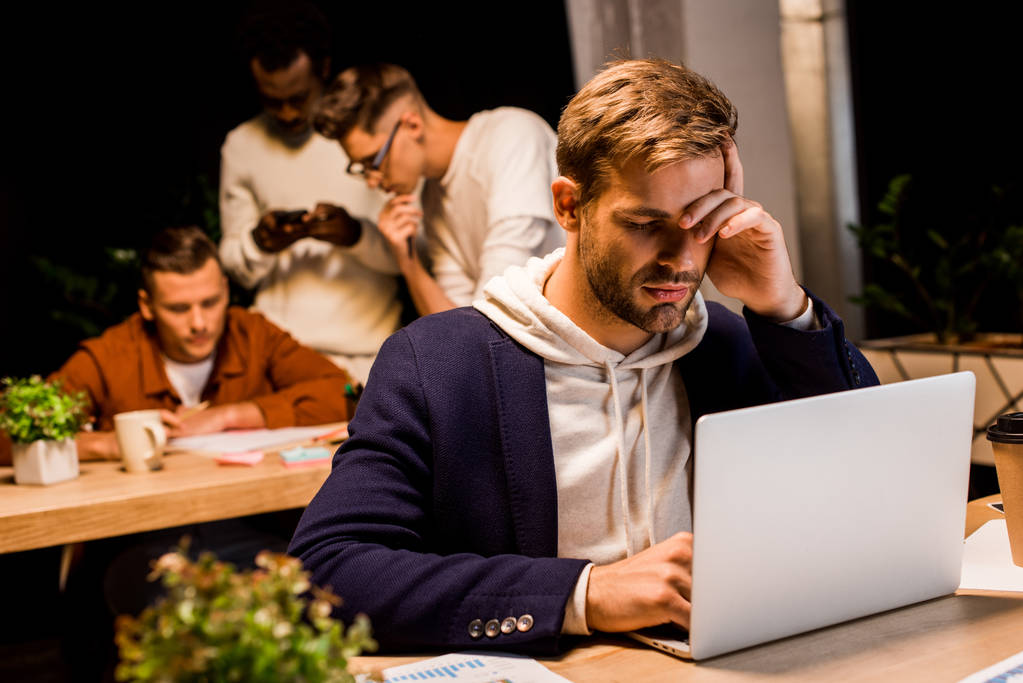 κουρασμένος επιχειρηματίας που χρησιμοποιεί φορητό υπολογιστή ενώ εργάζεται στο γραφείο τη νύχτα κοντά σε πολυπολιτισμικούς συναδέλφους - Φωτογραφία, εικόνα