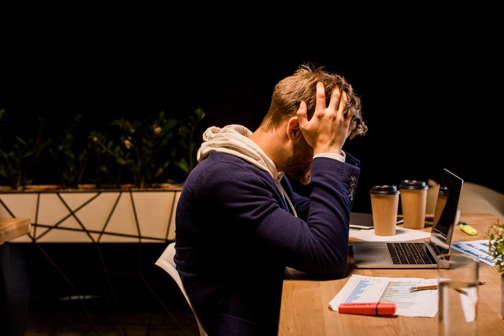 giovane uomo d'affari esausto mani vuote sulla testa mentre seduto di notte in ufficio vicino a laptop e bicchieri di carta
  - Foto, immagini