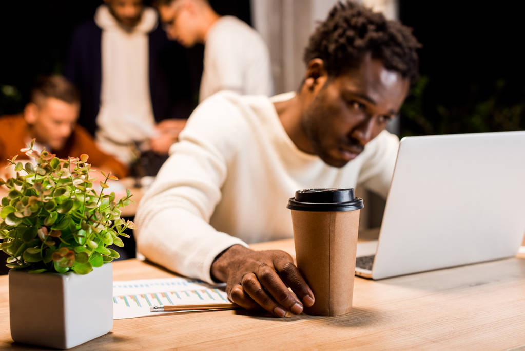 επιλεκτική εστίαση της εξαντλημένης Αφρικής Αμερικανός επιχειρηματίας κρατώντας καφέ για να πάει, ενώ εργάζονται τη νύχτα στο γραφείο κοντά σε συναδέλφους - Φωτογραφία, εικόνα