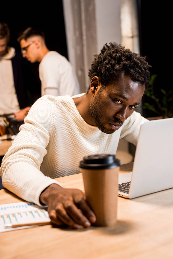 wybiórcze skupienie zmęczonego afrykańskiego biznesmena trzymającego kawę na wynos podczas pracy w biurze w pobliżu kolegów - Zdjęcie, obraz