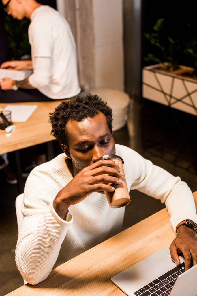 κουρασμένος αφροαμερικανός επιχειρηματίας πίνοντας καφέ για να πάει, ενώ εργάζονται το βράδυ στο γραφείο - Φωτογραφία, εικόνα