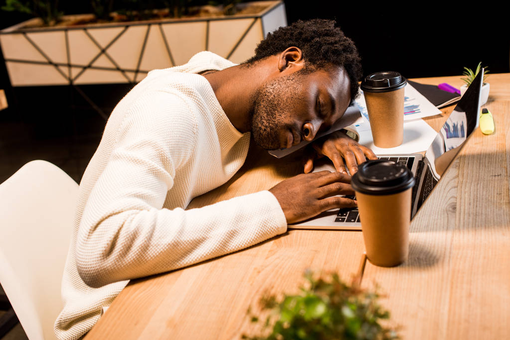 épuisé homme d'affaires afro-américain dormant sur le lieu de travail près d'un ordinateur portable et des tasses jetables la nuit au bureau
 - Photo, image