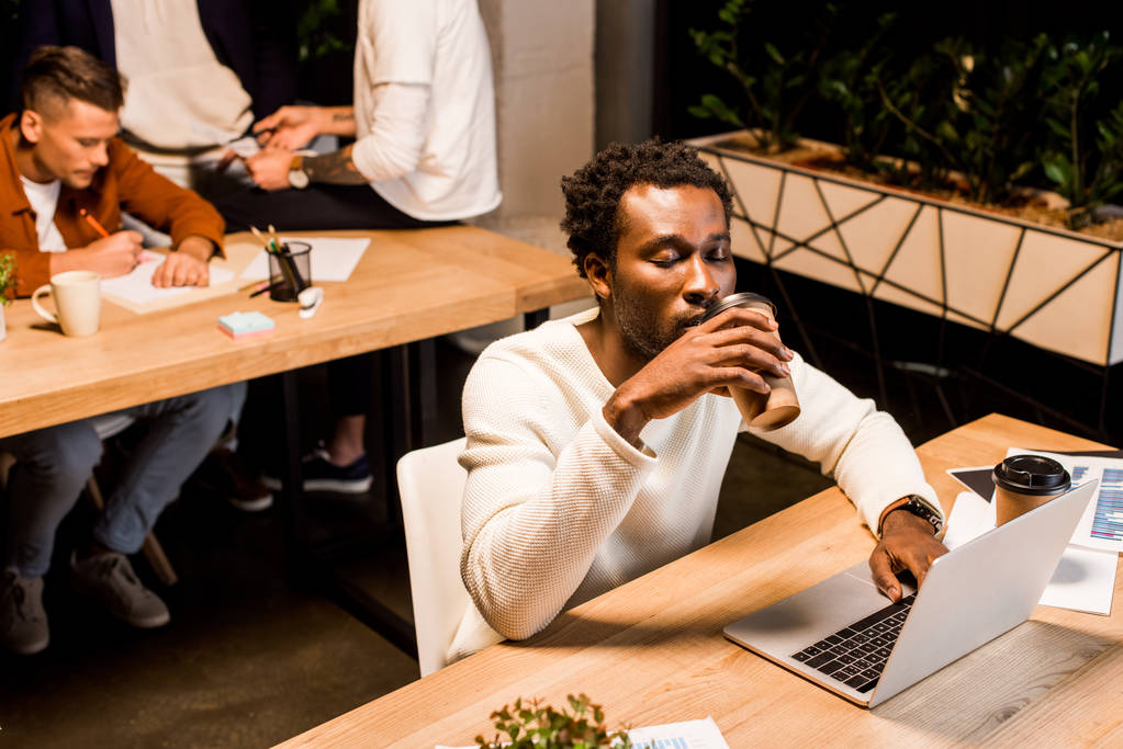 zmęczony afrykański amerykański biznesmen pijący kawę na wynos siedząc w pracy w biurze w pobliżu kolegów - Zdjęcie, obraz
