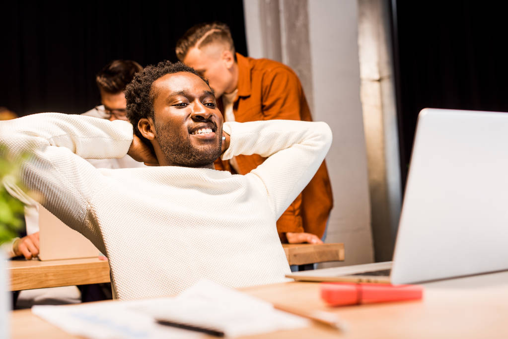 ευτυχισμένος Αφροαμερικάνος επιχειρηματίας που κοιτάζει το laptop ενώ εργάζεται τη νύχτα με συναδέλφους - Φωτογραφία, εικόνα