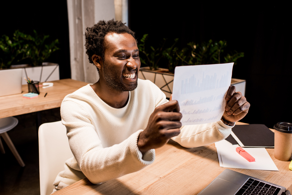 szczęśliwy afrykański amerykański biznesmen patrząc na papier podczas pracy w nocy w biurze - Zdjęcie, obraz