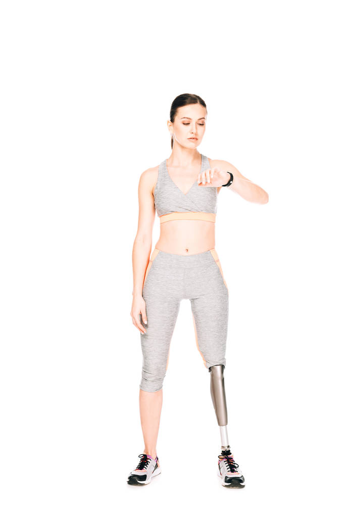 Ganzkörperansicht der Sportlerin mit Beinprothese beim Betrachten der Smartwatch isoliert auf Weiß - Foto, Bild