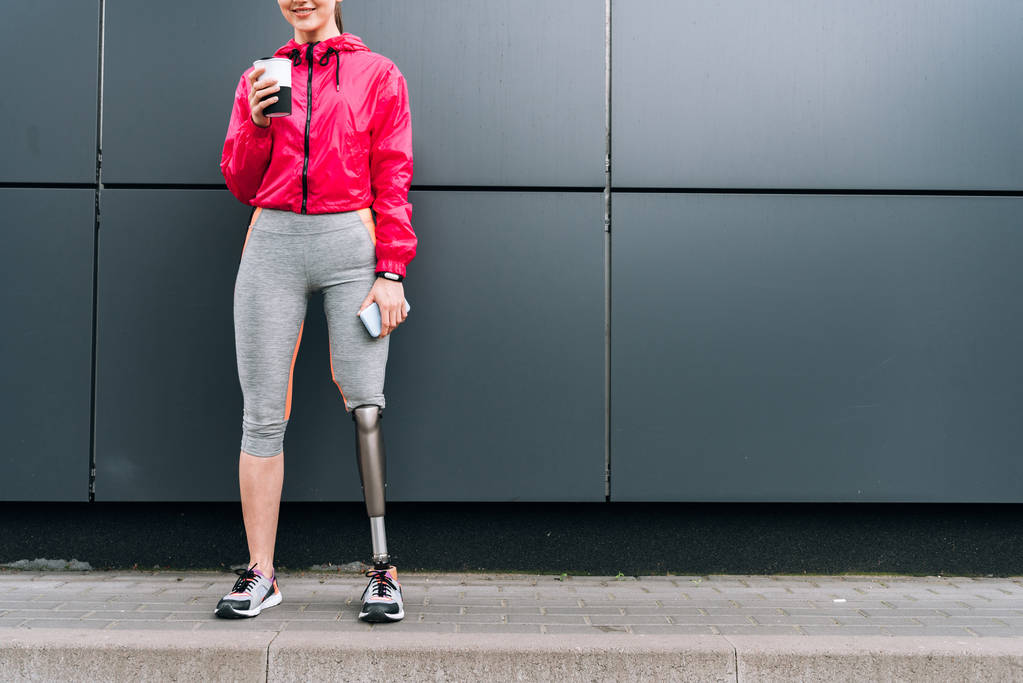 vista recortada de la sonriente deportista discapacitada sosteniendo taza y teléfono inteligente en la calle
 - Foto, imagen