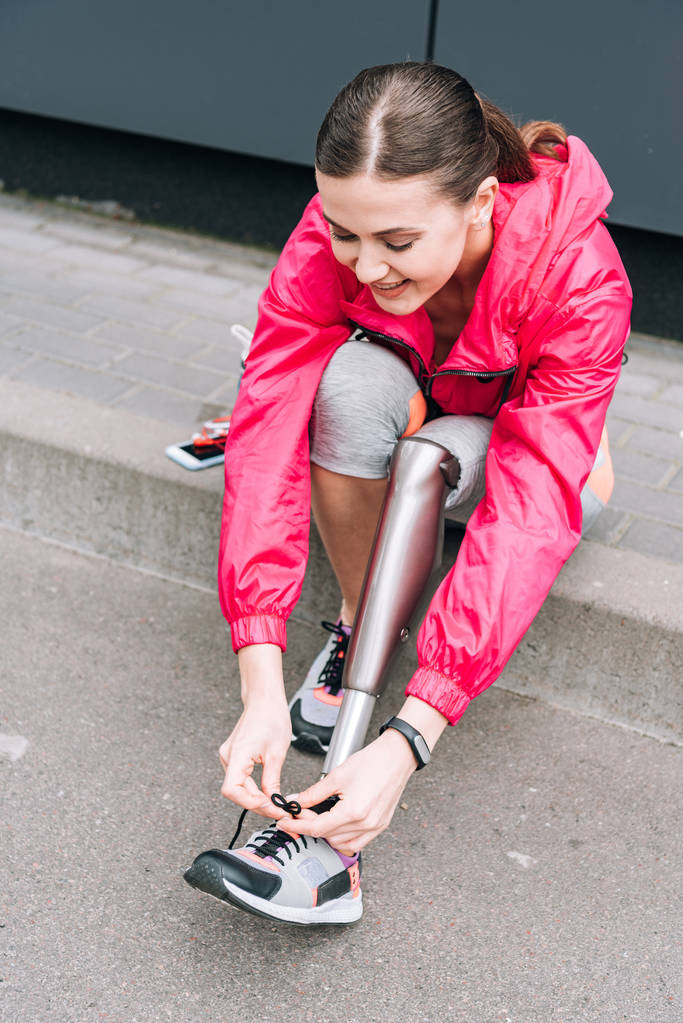 улыбающаяся спортсменка-инвалид с протезом, завязывающая шнурки на улице
 - Фото, изображение