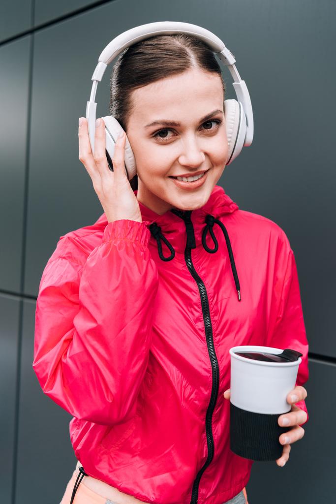 mosolygós sportos nő kezében csésze és zenét hallgat a fejhallgató az utcán - Fotó, kép