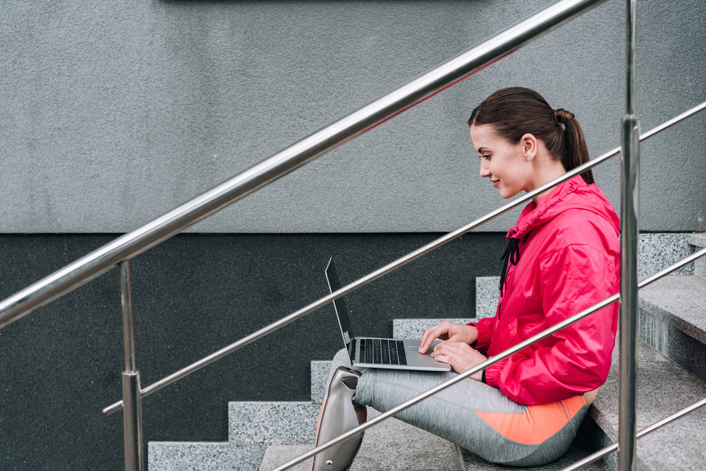 boční pohled na usmívající se zdravotně postižené sportovkyně pomocí notebooku při sezení na schodech na ulici - Fotografie, Obrázek