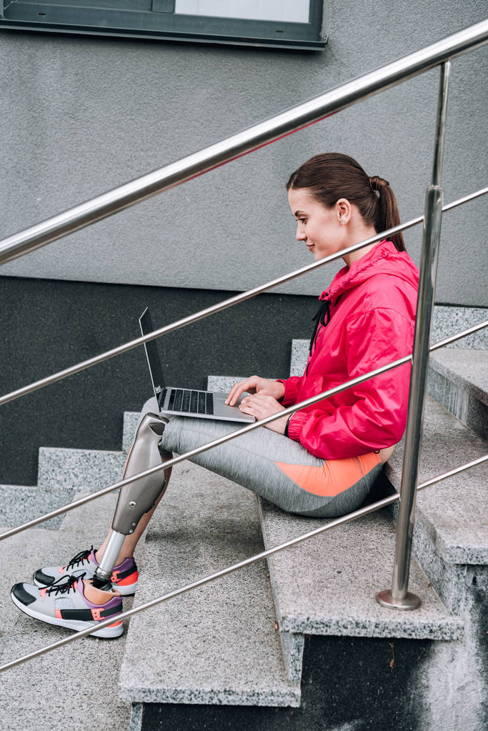 Sokakta merdivenlerde otururken dizüstü bilgisayar kullanan engelli sporcunun yan görüntüsü - Fotoğraf, Görsel