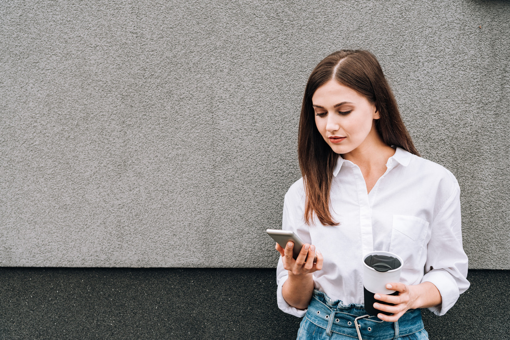 Güzel genç bir kadın elinde akıllı telefon ve kupayla caddede - Fotoğraf, Görsel