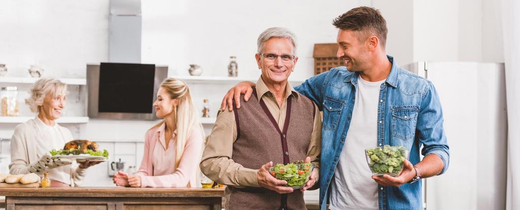 панорамный снимок улыбающихся дедушки и папы, обнимающих и держащих миски с салатом и брокколи в День Благодарения
  - Фото, изображение