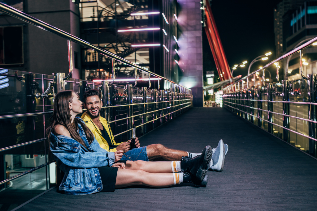 vista lateral de novio con botella y novia sonriente sentado en la ciudad de la noche
  - Foto, Imagen