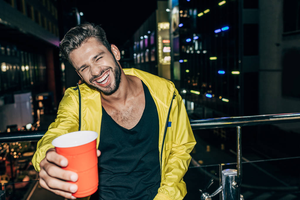 Красивый мужчина в жёлтой куртке улыбается и держит пластиковую чашку в ночном городе
  - Фото, изображение