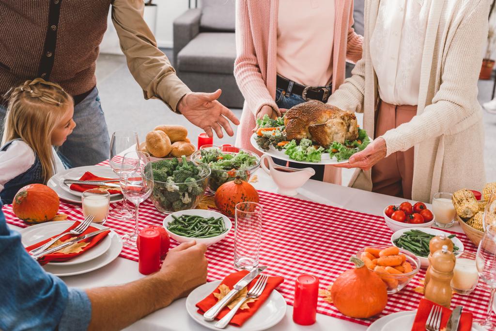 bijgesneden uitzicht op familie zitten aan tafel en moeder met oma holding plaat met kalkoen in Thanksgiving dag      - Foto, afbeelding