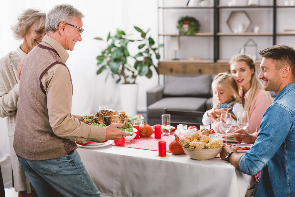 familiares sentados a la mesa y el abuelo sosteniendo el plato con pavo en el día de Acción de Gracias
       - Foto, imagen