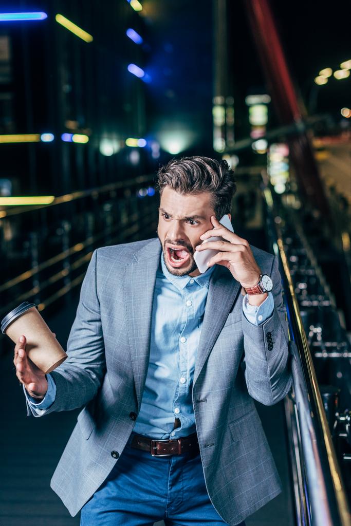 dühös jóképű üzletember formális kopás beszél a smartphone és a gazdaság papírpohár éjszakai városban   - Fotó, kép