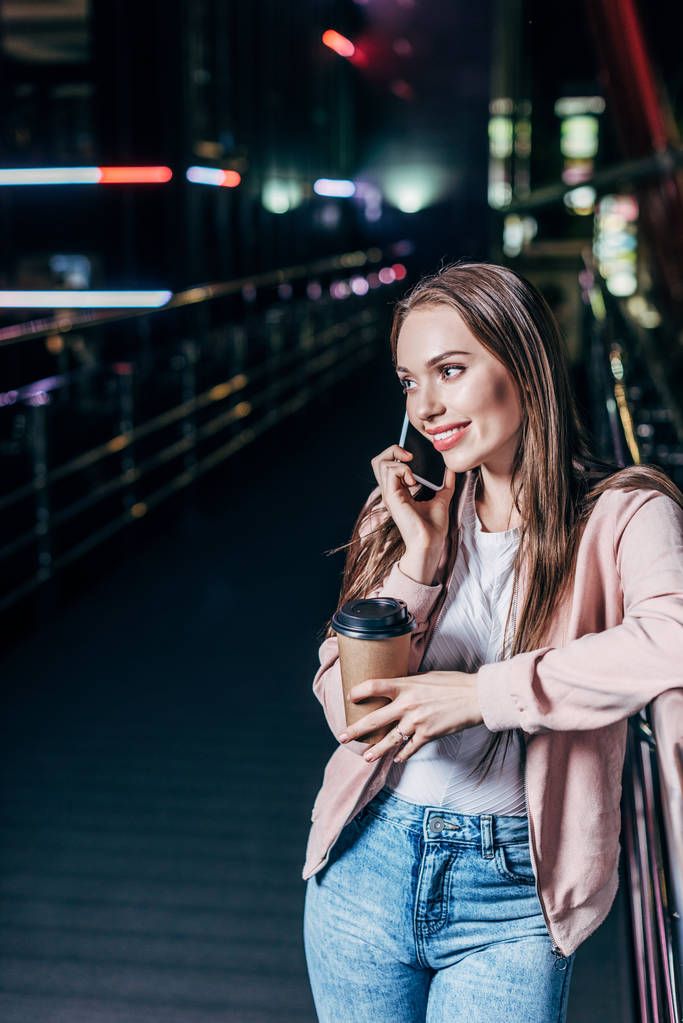attraktive Frau in pinkfarbener Jacke, die mit dem Smartphone spricht und in der Nacht einen Pappbecher in der Hand hält  - Foto, Bild