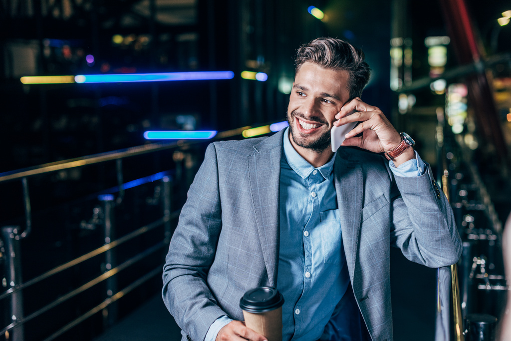 guapo hombre de negocios en ropa formal hablando en el teléfono inteligente y sosteniendo taza de papel en la ciudad de la noche
   - Foto, Imagen