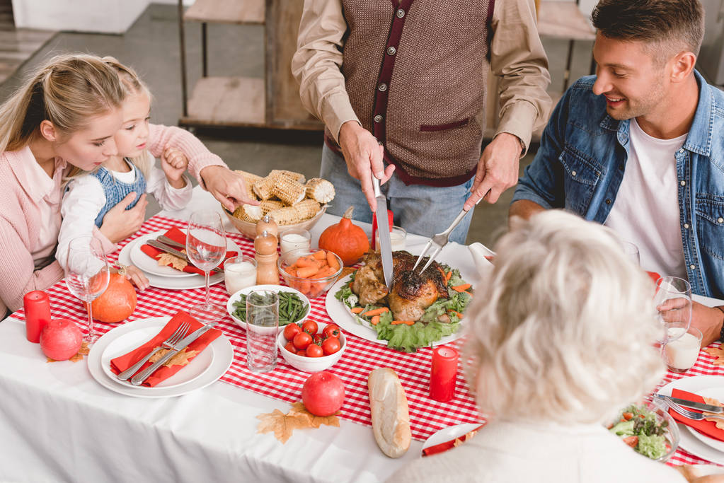 abgeschnittene Ansicht von Familienmitgliedern, die am Tisch sitzen und Großvater schneidet schmackhaften Truthahn zum Erntedank       - Foto, Bild