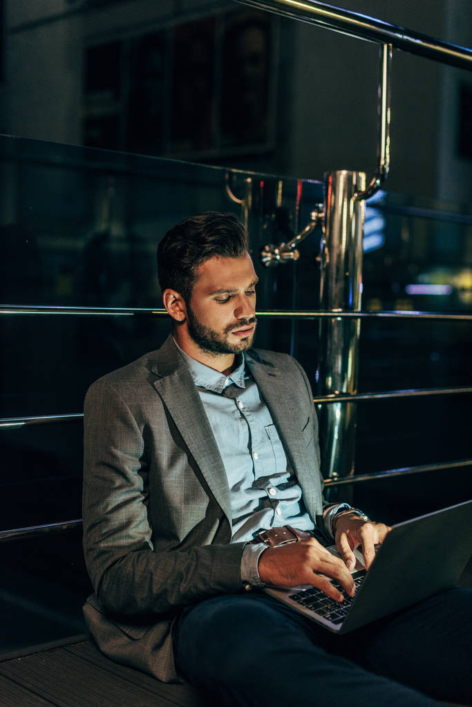 όμορφος επιχειρηματίας στην επίσημη ένδυση καθιστός και χρησιμοποιώντας το laptop στη νυχτερινή πόλη   - Φωτογραφία, εικόνα