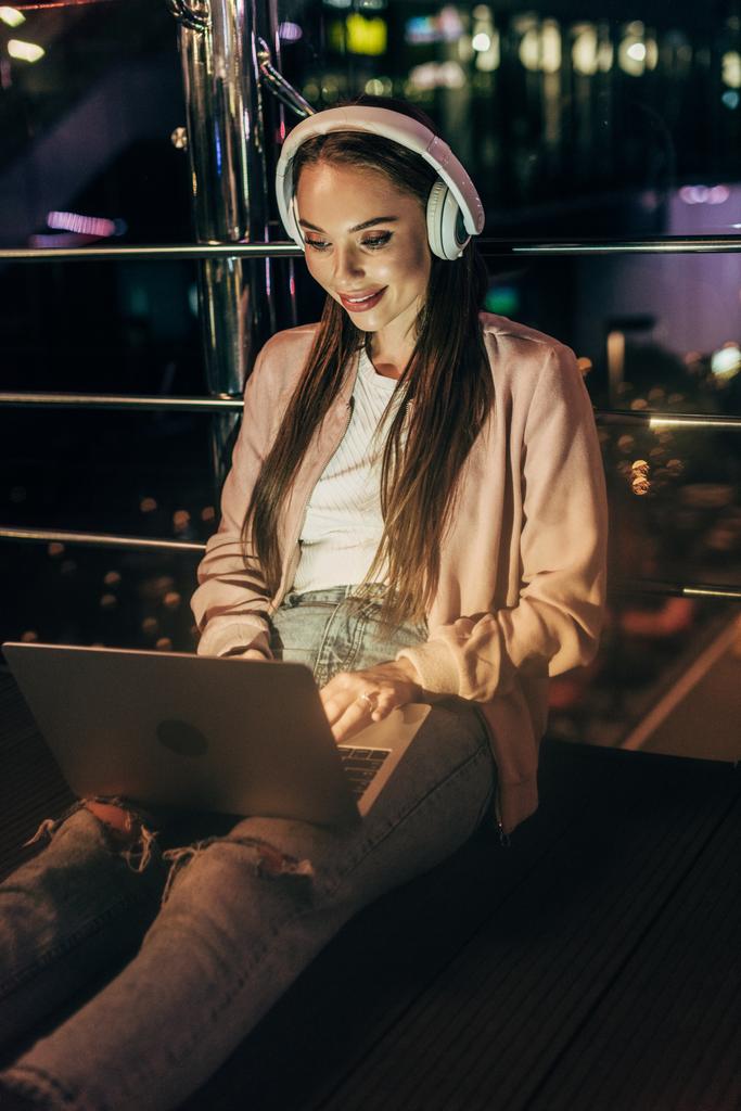 donna sorridente in giacca rosa con cuffie utilizzando laptop in città di notte
   - Foto, immagini