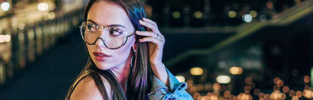 tiro panorâmico de mulher atraente em óculos olhando para longe na cidade noturna
   - Foto, Imagem